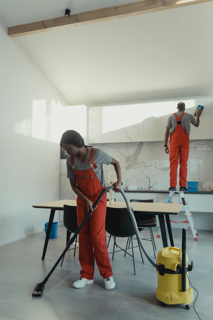 Two professional cleaners from Bayswater Cleaner are conducting surface cleaning and deep cleaning in a modern kitchen. One cleaner is using a vacuum or carpet cleaner on the wooden or laminate flooring, ensuring thorough hygiene and dust removal. The other cleaner is standing on a step ladder, wiping or polishing a large wall-mounted marble or stone backsplash with a cloth or spray. The kitchen features smooth, glossy countertops, a white marble or ceramic backsplash with subtle veining, and a light-colored ceiling with wooden beam accents. Natural light streams into the room through windows, illuminating the clean and well-maintained space. The setting highlights the company's expertise in domestic cleaning and sanitisation, ensuring surfaces are spotless and hygienic, consistent with the services described on Bayswater Road end of tenancy cleaning experts W2 page at bayswatercleaner.co.uk.
