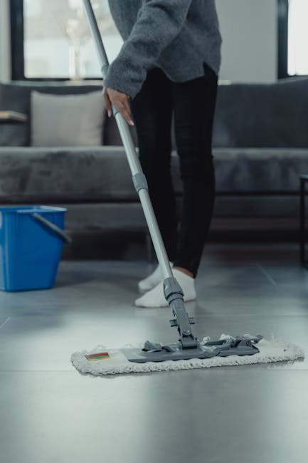 A person wearing black trousers and white sneakers is cleaning a dark tiled living room floor using a flat mop with a white microfiber head. The person is positioned slightly to the left, with their hand gripping the handle of the mop, which is being moved across the surface. In the background, there is a grey sofa with cushions, a black side table, and large windows allowing natural light to fill the space. To the left of the person, a blue cleaning bucket is placed on the floor. The clean, shiny floor indicates a thorough surface cleaning process, showcasing the professional cleaning services of Bayswater Cleaner for domestic or commercial properties in the W2 area, emphasizing surface cleaning and sanitisation.
