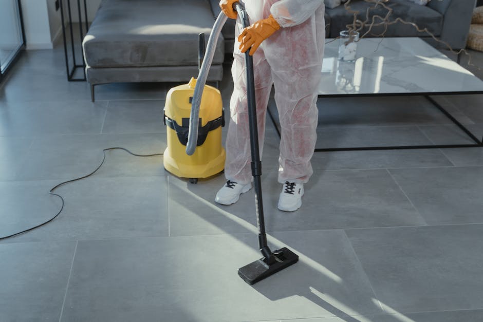 Two professional cleaners from Bayswater Cleaner are conducting surface cleaning and deep cleaning in a modern kitchen. One cleaner is using a vacuum or carpet cleaner on the wooden or laminate flooring, ensuring thorough hygiene and dust removal. The other cleaner is standing on a step ladder, wiping or polishing a large wall-mounted marble or stone backsplash with a cloth or spray. The kitchen features smooth, glossy countertops, a white marble or ceramic backsplash with subtle veining, and a light-colored ceiling with wooden beam accents. Natural light streams into the room through windows, illuminating the clean and well-maintained space. The setting highlights the company's expertise in domestic cleaning and sanitisation, ensuring surfaces are spotless and hygienic, consistent with the services described on Bayswater Road end of tenancy cleaning experts W2 page at bayswatercleaner.co.uk.