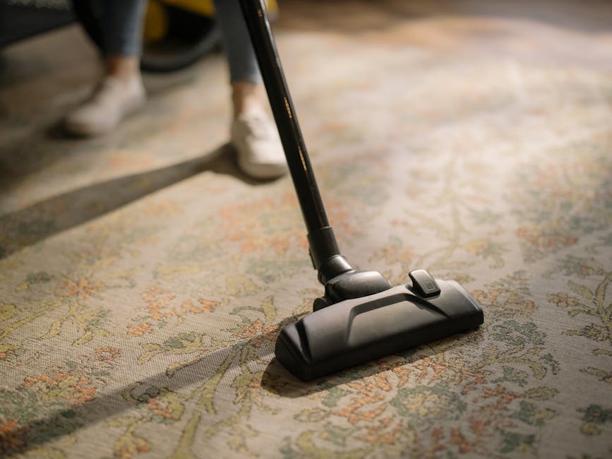 A child using a vacuum cleaner on a patterned, beige and pastel floral area rug in a room with wooden flooring visible around the edges. The vacuum cleaner has a yellow body with a black hose and a me