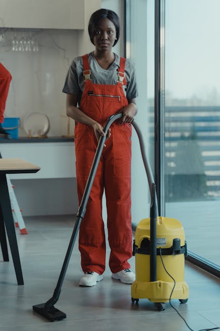 A professional cleaner from Bayswater Cleaner stands indoors on a light grey tiled floor, operating a yellow commercial vacuum cleaner. She is wearing a grey t-shirt and bright orange overalls, with b