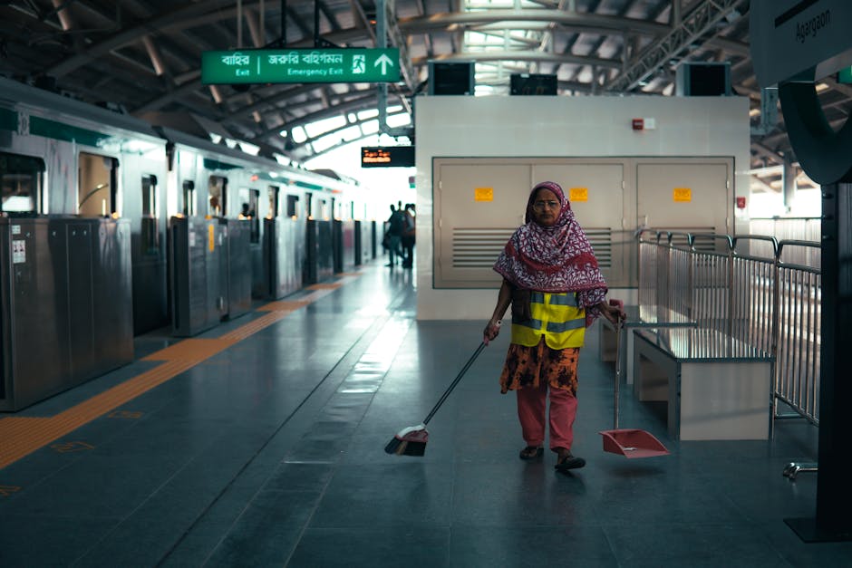 A professional cleaner from Bayswater Cleaner stands indoors on a light grey tiled floor, operating a yellow commercial vacuum cleaner. She is wearing a grey t-shirt and bright orange overalls, with b